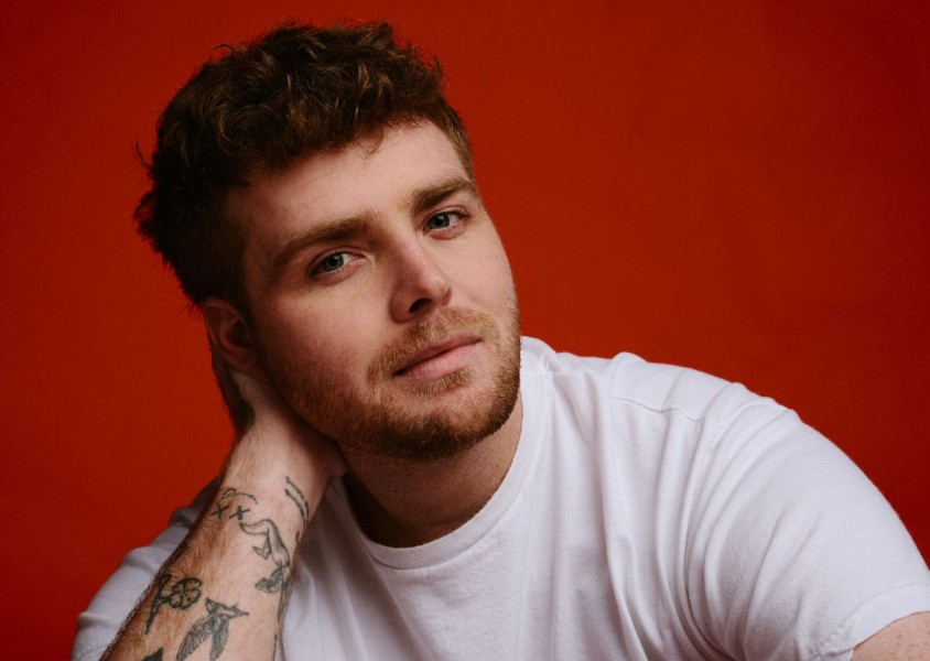 A man in a white tshirt with short brown hair in front of a red backgroundx